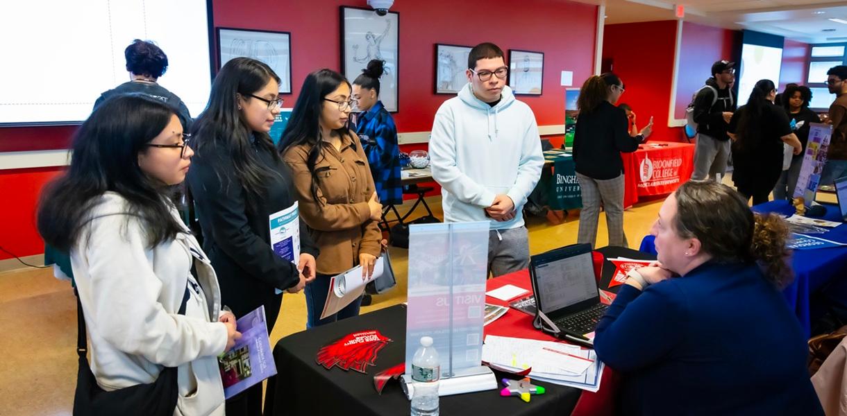 Students speak with a college representative at an information table during a transfer fair, while other college tables and attendees are visible in the background.