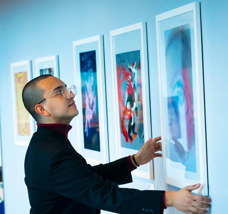 An artist adjusts a framed artwork displayed on a gallery wall featuring a series of colorful, framed pieces in the HCCC Foundation Art Collection.