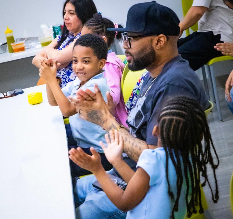 Adults and children seated together around a classroom-style table, participating in a group activity in a bright community space with refreshments on a nearby counter.