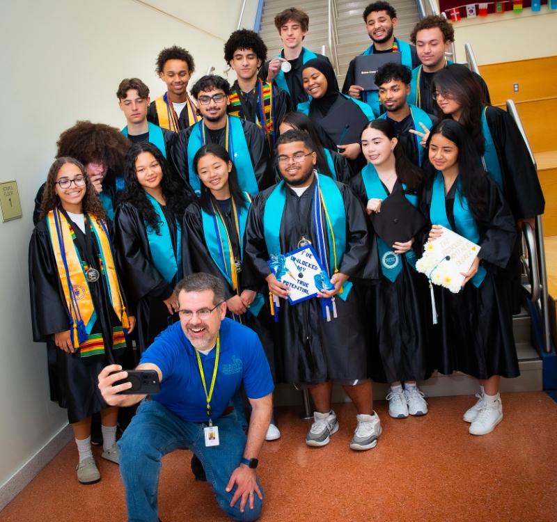 A group of graduating students wearing caps, gowns, honor cords, and stoles gather on a school staircase for a photo while a staff member kneels in front, taking a selfie with a smartphone. One graduate holds a decorated mortarboard displaying the message, “Proud of What We’ve Unlocked Our Potential.”