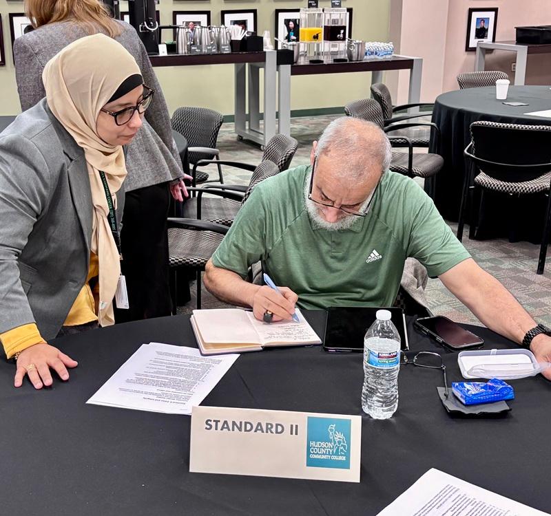 Two meeting participants at a conference table, with one person writing notes in a notebook while another stands nearby reviewing documents. A table sign reads “STANDARD II” and includes the Hudson County Community College logo.