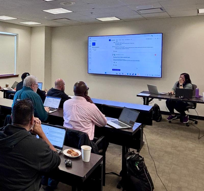 Participants seated in a classroom-style training session using laptops while viewing a presentation displayed on a large screen at the front of the room.
