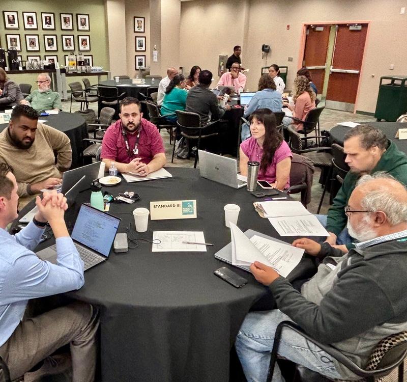 Participants seated around a round table reviewing documents and using laptops during a group discussion. A table sign reads “STANDARD III,” with additional discussion groups visible in the conference room.