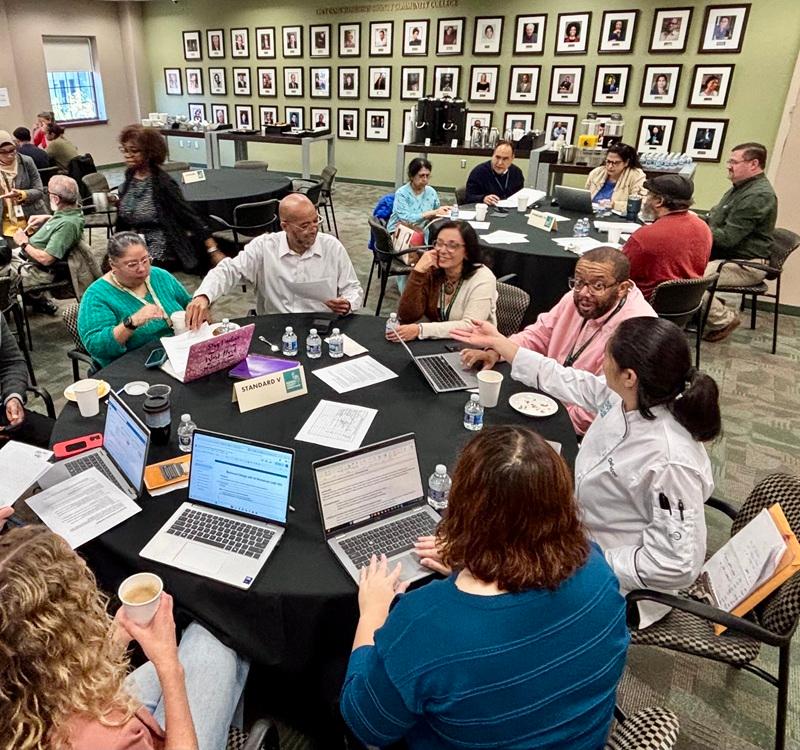 Participants seated at round tables during a meeting, using laptops and reviewing documents in a conference room. A sign on the foreground table reads “STANDARD V.”