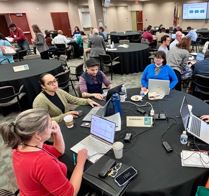 Participants seated around a round table using laptops and discussing documents during a collaborative meeting. A sign on the table reads “STANDARD I.”