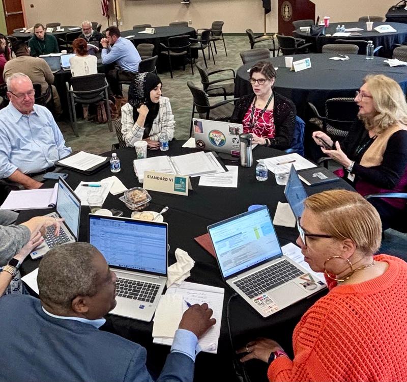 Participants seated around a round table with laptops, documents, and water bottles engaged in a group discussion during a meeting. A table sign reads “STANDARD VI.”