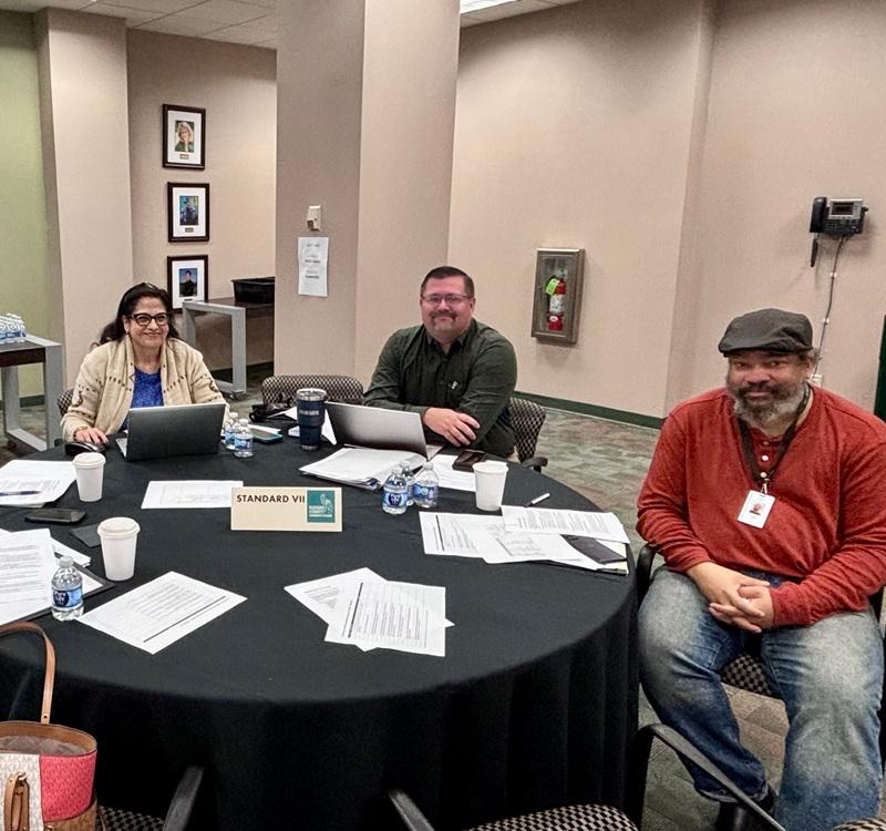 Three people seated around a conference table with laptops, documents, and drinks during a meeting. A sign on the table reads “STANDARD VII.”