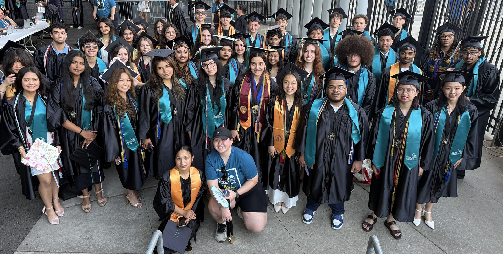 Early College Graduates at Commencement A large group of diverse college graduates in black academic regalia pose for a group photo outdoors at a commencement ceremony. The graduates wear matching teal and black stoles over their gowns, with several individuals also wearing additional honor cords and medals. In the center foreground, two individuals are kneeling: one in a black gown with an orange stole, and another wearing a blue casual t-shirt and shorts. The background shows an outdoor venue with concrete flooring and a dark metal fence.